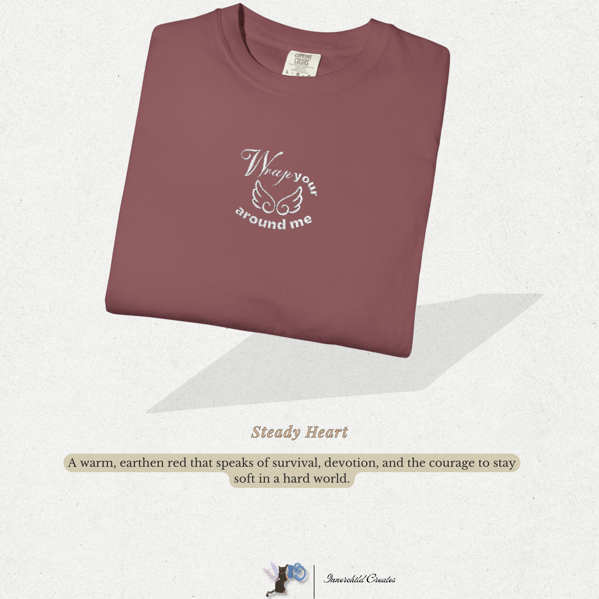 Folded maroon shirt with text on a light gray background. Brick, maroon, folded shirt with white embroidery with white wings