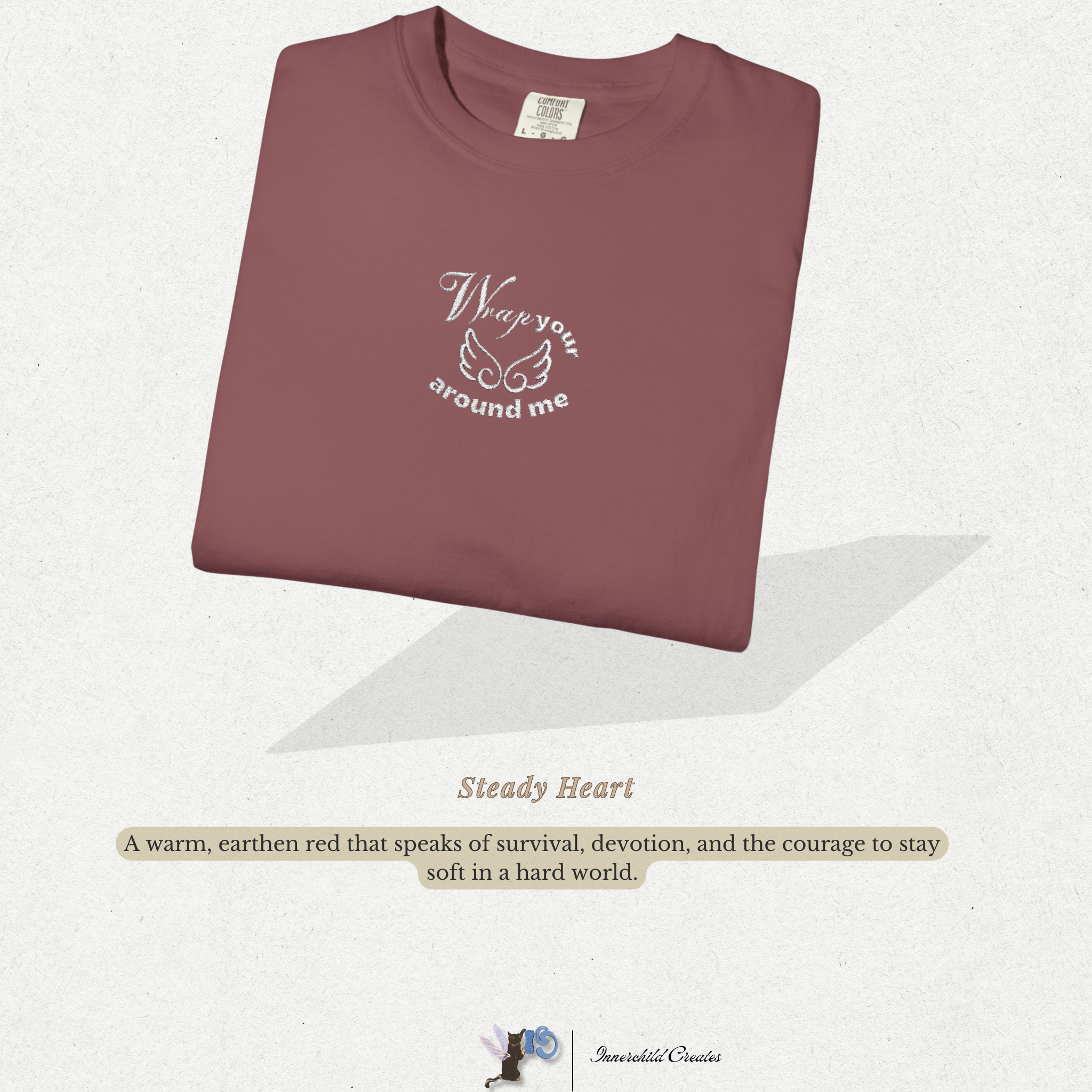 Folded maroon shirt with text on a light gray background. Brick, maroon, folded shirt with white embroidery with white wings