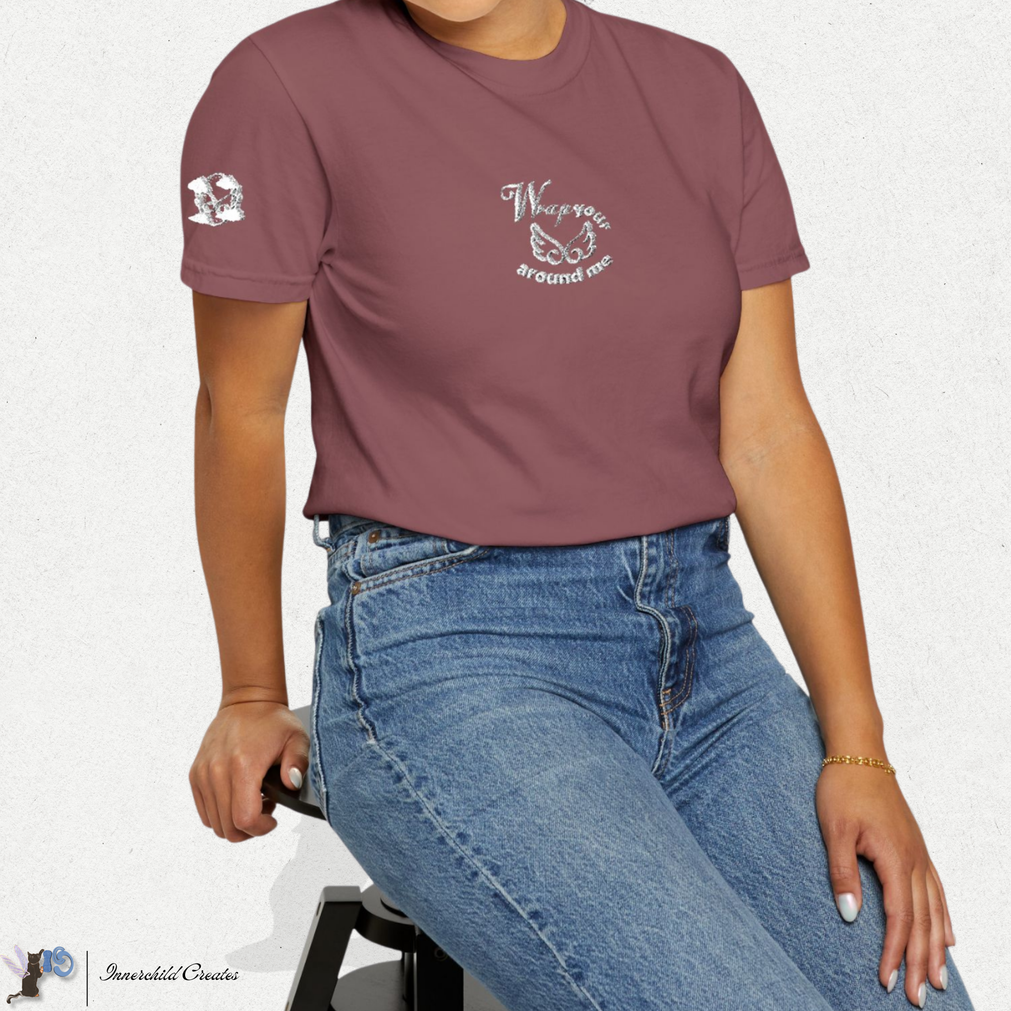 Person wearing a maroon t-shirt with text and graphics, sitting on a white background. Brick t-shirt. White embroidery wings & clouds. 