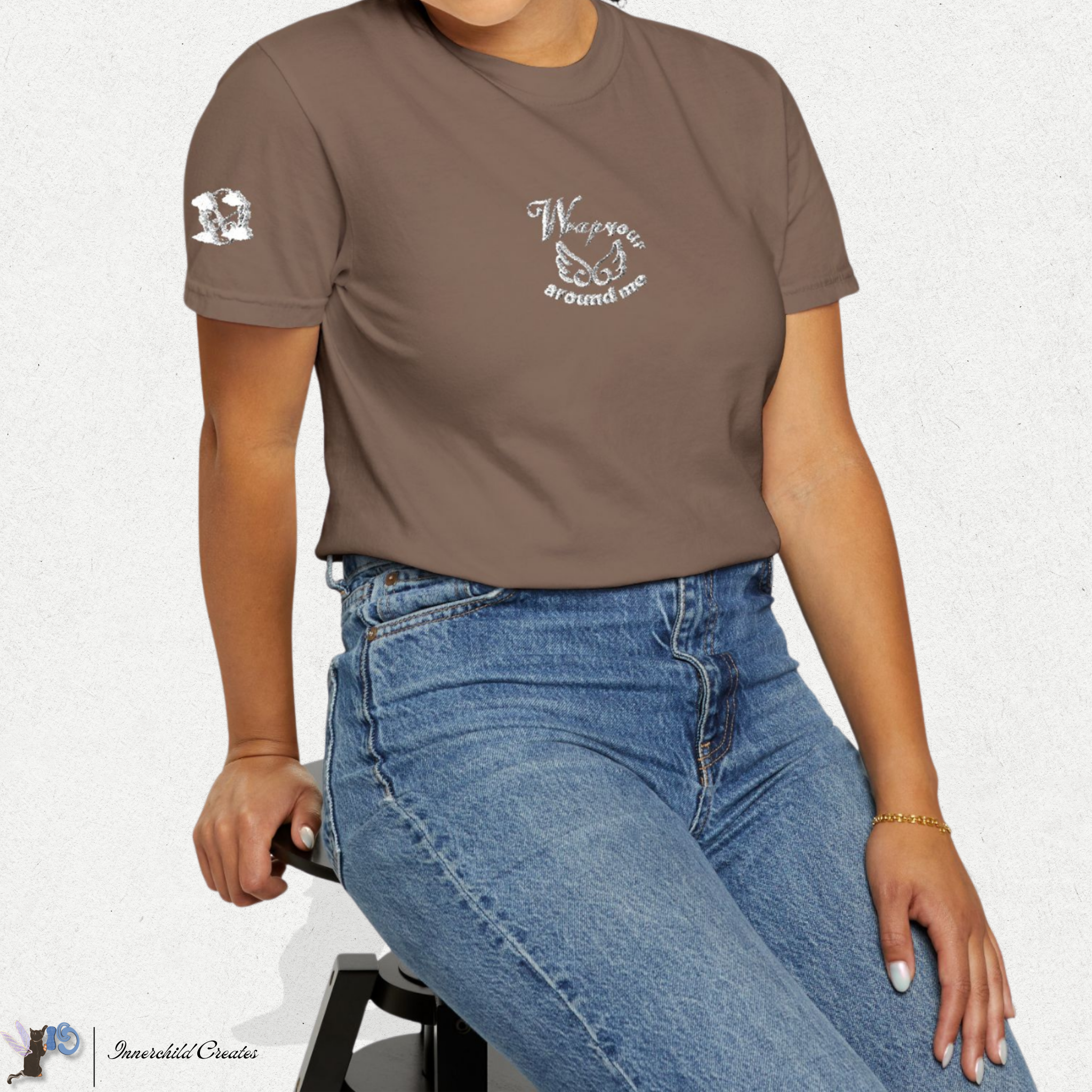 Person wearing a brown, espresso t-shirt with text and graphics, sitting on a white background. White embroidered of wings, clouds, and text.