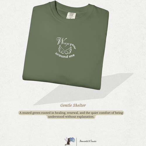 Folded green shirt with text on a light gray background. Sage, green folded shirt with white wings and cloud embroidery. 